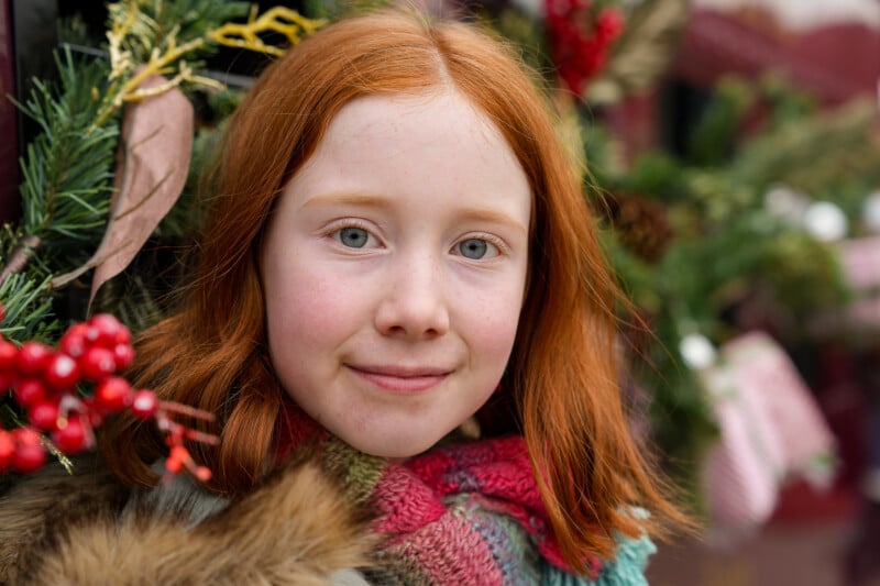 A young girl with long red hair and blue eyes smiles softly. She is wearing a colorful scarf and standing in front of festive greenery with red berries and pinecones. The background is blurred.