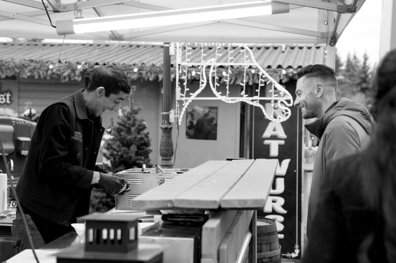 A smiling vendor prepares food at an outdoor market stall while a customer stands across the counter, happily engaging in conversation. The scene is lively and casual, with festive lights in the background.