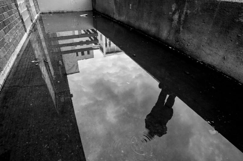 A black-and-white photo shows a puddle in an alleyway reflecting buildings, clouds, and a person standing nearby, whose silhouette is visible in the water. The walls and ground are gritty and urban.