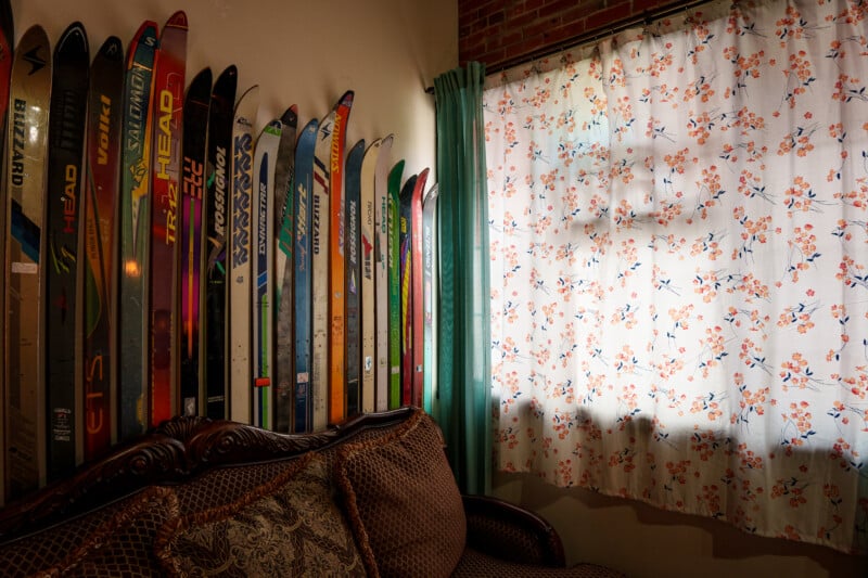 A collection of colorful vintage skis is lined up against a wall behind a brown patterned sofa, next to a window with sheer floral curtains and green drapes. Sunlight streams in, casting shadows on the curtain.