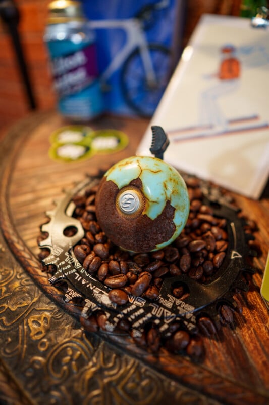 A rusty, blue-green metal gear shift sits on a bed of coffee beans, arranged inside a bicycle chainring on a carved wooden surface. Blurred background includes a can, a bike image, and other colorful objects.