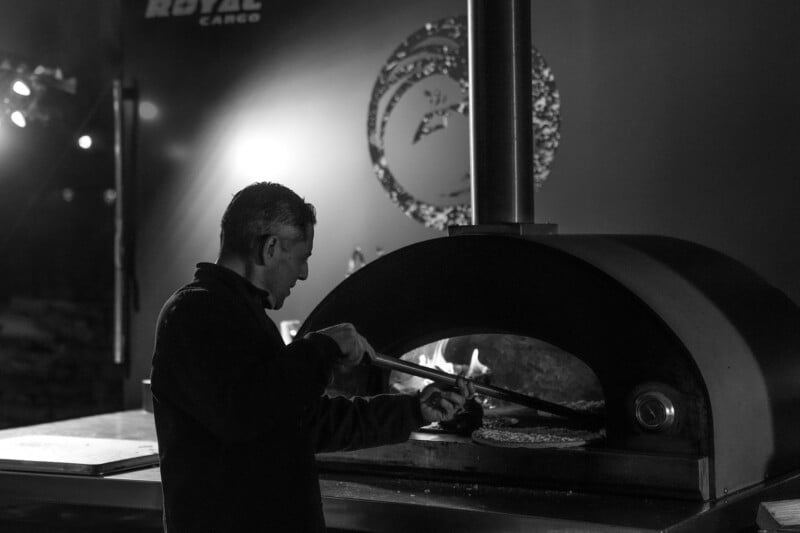 A person uses a long-handled tool to bake a pizza in a wood-fired oven. Flames are visible inside the oven, and the scene is lit with dramatic lighting in black and white.