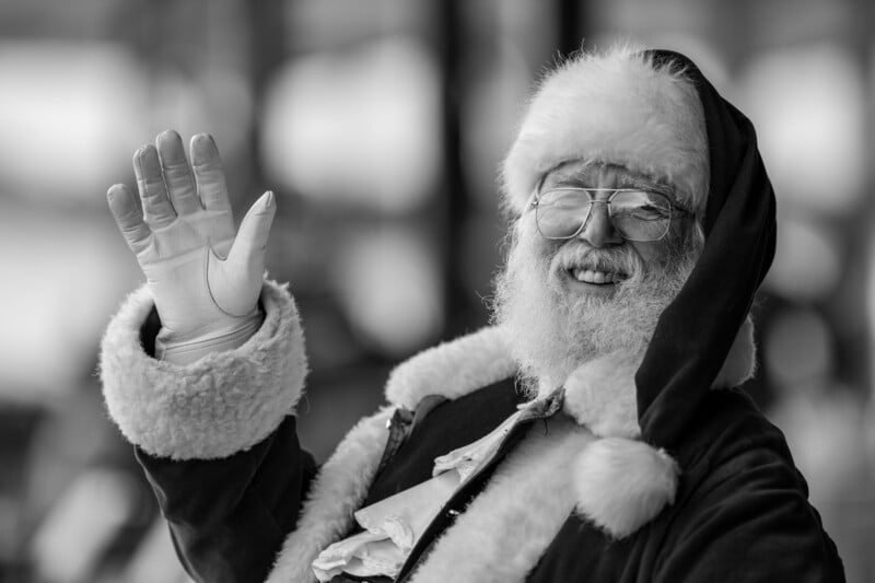A person dressed as Santa Claus, with a white beard and glasses, smiles and waves while wearing a fur-trimmed hat and coat. The image is in black and white.