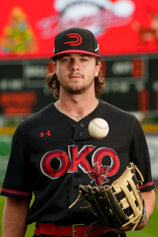 A baseball player in a black and red uniform labeled "OKO" stands holding a glove and tossing a baseball. The background shows a scoreboard and a large red sign with blurred graphics.