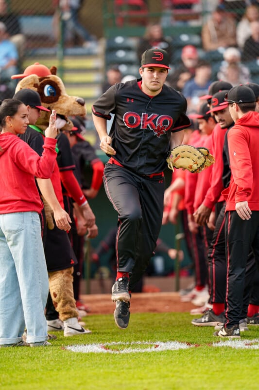 A baseball player in a black OKO uniform and cap runs onto the field through a line of cheering teammates and a mascot, preparing for a game. Spectators watch from the stands in the background.