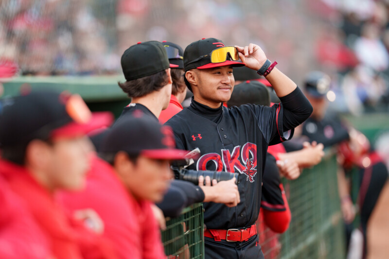 A baseball player in a black and red uniform stands by the dugout, smiling and adjusting his sunglasses, while teammates in matching uniforms watch the game beside him.