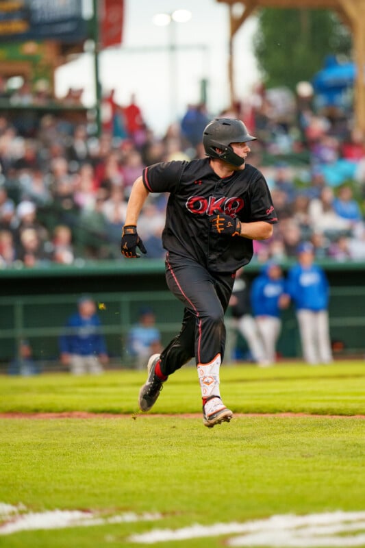 A baseball player in a black uniform and helmet runs on a grassy field during a game, with a blurred crowd and other players in blue uniforms in the background.