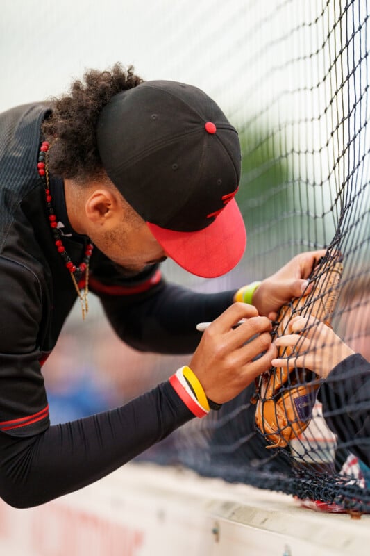 A baseball player wearing a black and red uniform signs a baseball through a net for a fan, leaning forward with a marker in hand.