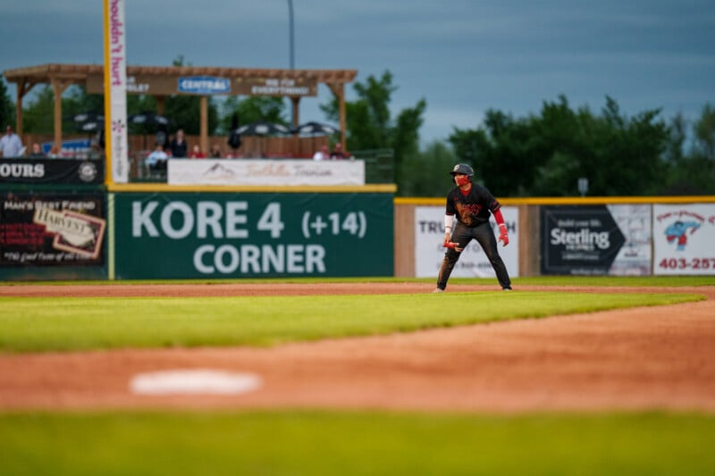 A baseball player in a black uniform stands ready on a base with a green outfield and advertising signs in the background under a cloudy sky.
