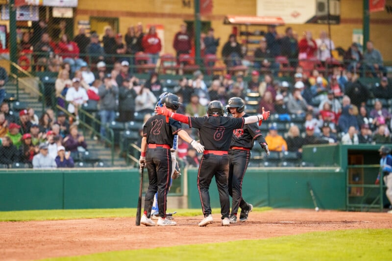 Three baseball players in black uniforms celebrate at home plate, arms outstretched, after scoring a run. Spectators watch from the stands in the background. The atmosphere appears energetic and lively.