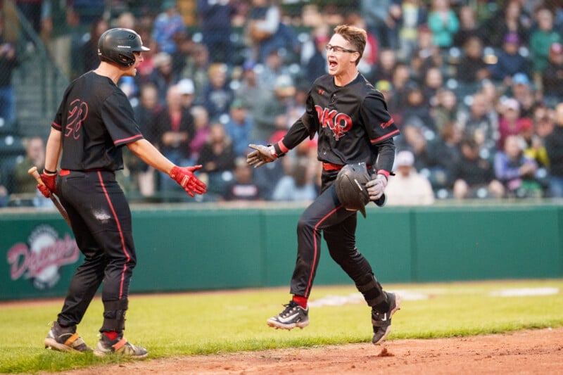 A baseball player in a black uniform jumps in excitement while holding his helmet, as a teammate reaches out for a high-five on a baseball field with a crowd in the background.