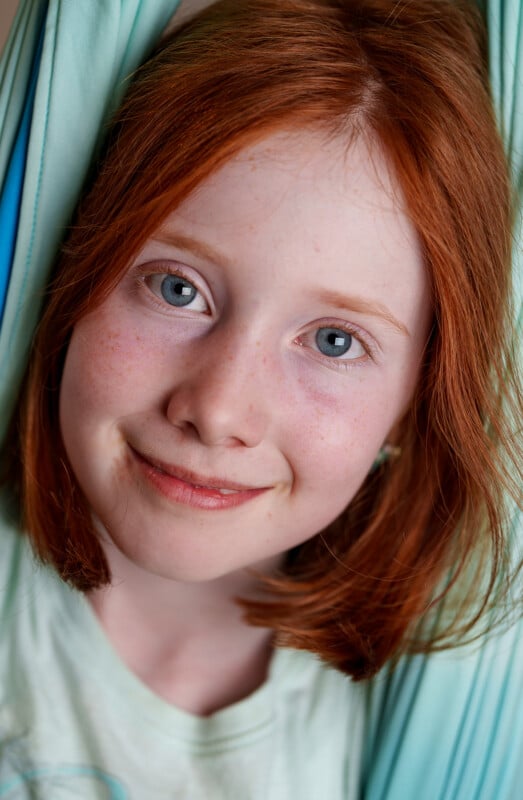 A close-up of a smiling child with light skin, blue eyes, and shoulder-length red hair, wearing a light-colored shirt, framed by light blue fabric on both sides.
