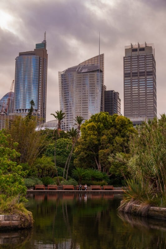 Tall modern skyscrapers rise above lush green trees and a reflective pond, with benches and people near the water in an urban park, under a cloudy sky.