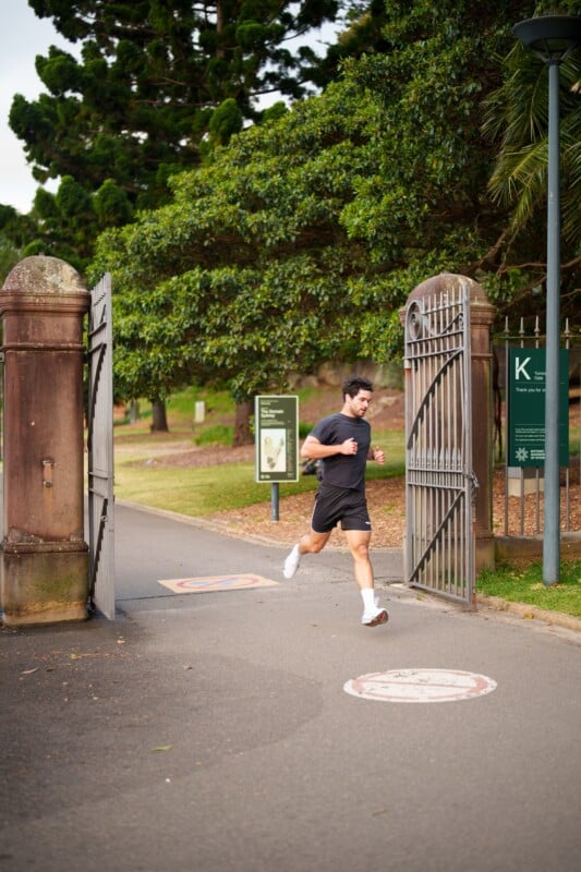 A man in black athletic clothes jogs through open iron gates in a park surrounded by green trees and signs, on a paved path with a pedestrian symbol painted on the ground.