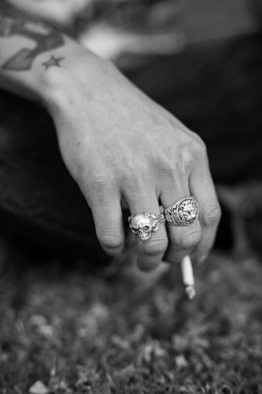 A close-up of a hand with two large rings—one shaped like a skull—holding a cigarette. The forearm shows a star tattoo. The background is blurred grass. The image is in black and white.