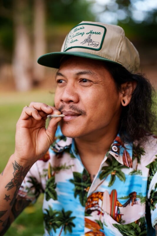 A man wearing a tropical print shirt and a beige cap with a patch sits outdoors, holding a rolled cigarette or joint to his mouth. He has long hair, a mustache, and tattoos on his arm. Greenery is blurred in the background.