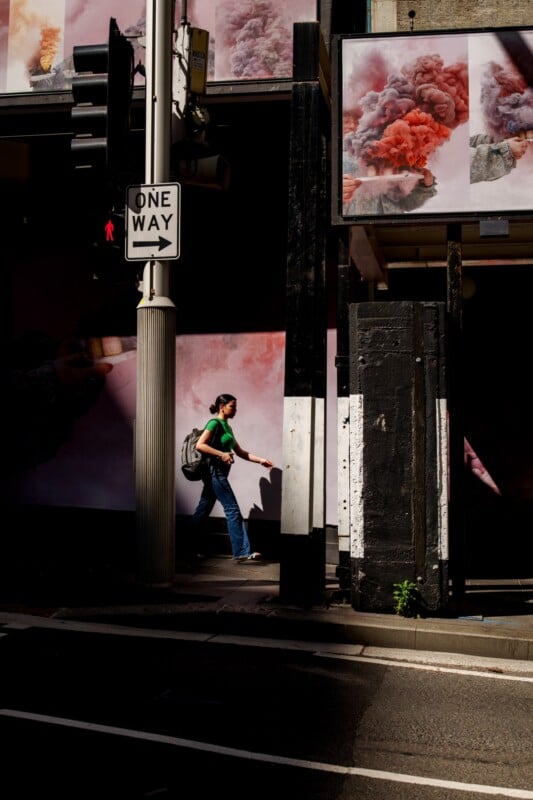 A person with a backpack walks on a city sidewalk, passing under a one-way street sign and in front of posters showing colorful smoke. Shadows and sunlight create dramatic contrasts on the scene.