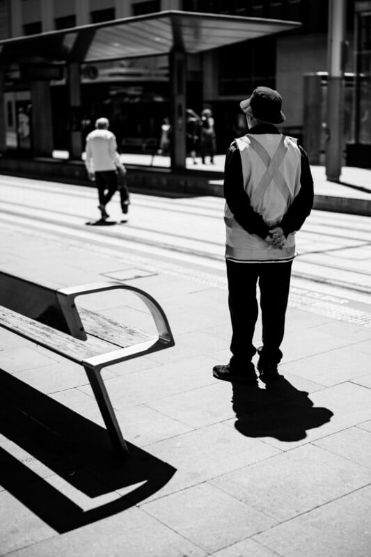 A person wearing a reflective vest and hat stands on a city sidewalk with hands behind their back; in the background, another person walks away near a tram stop on a sunny day.