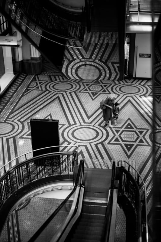 A person and a child walk across an elaborate, geometric tiled floor in a shopping mall, viewed from above near an escalator. The scene is in black and white.