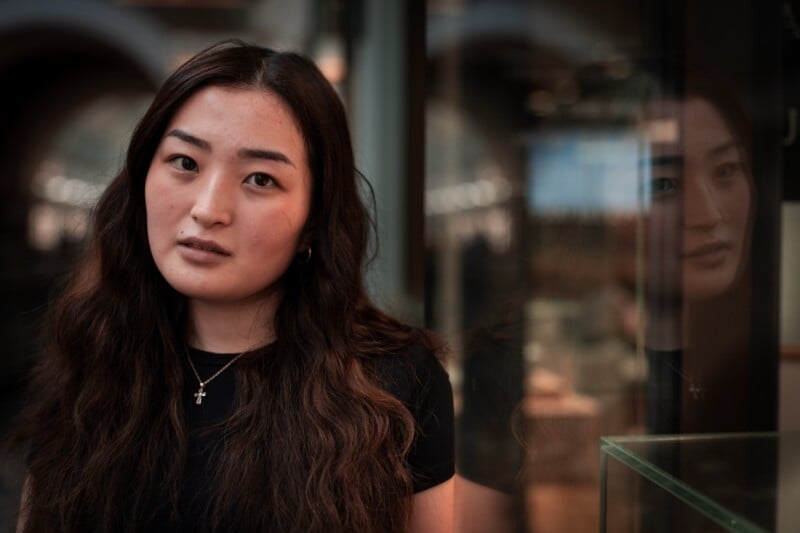 A woman with long wavy hair wearing a black top and a cross necklace stands indoors near a reflective glass surface, showing her face and her reflection, with a soft, blurred background.
