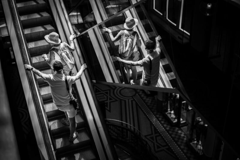 A person wearing a hat and backpack rides an escalator, reaching toward a mirrored wall reflecting their image in black and white, creating a surreal effect.