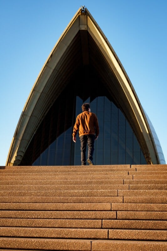 A person in a brown jacket walks up the steps toward the Sydney Opera House, with its iconic pointed roof and glass windows visible under a clear blue sky.