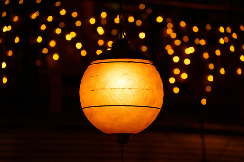 A glowing round lantern hangs in the foreground, illuminated with a warm golden light. In the blurry background, many small yellow lights create a festive, bokeh effect.