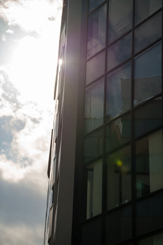 A modern glass building is seen from a low angle with sunlight shining through clouds, reflecting off the windows and creating a lens flare effect.