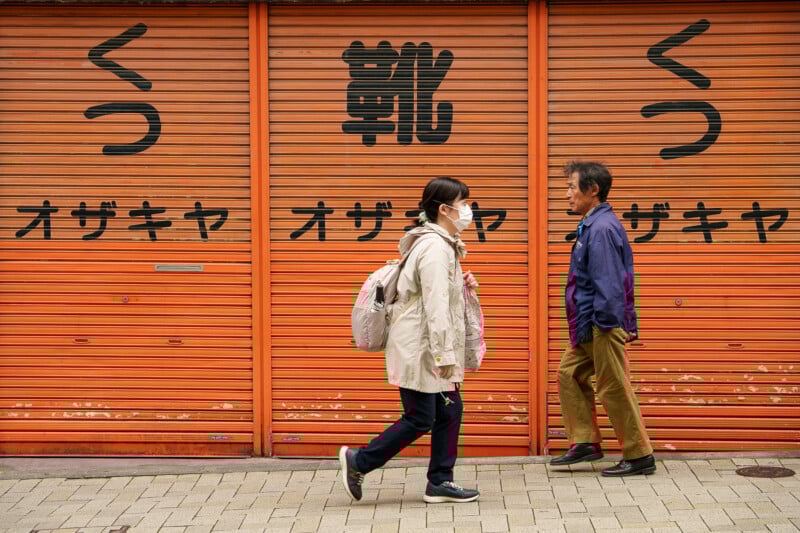 A woman wearing a mask and backpack walks past an older man in front of orange metal shutters with Japanese writing on them. Both are walking in opposite directions on a paved sidewalk.