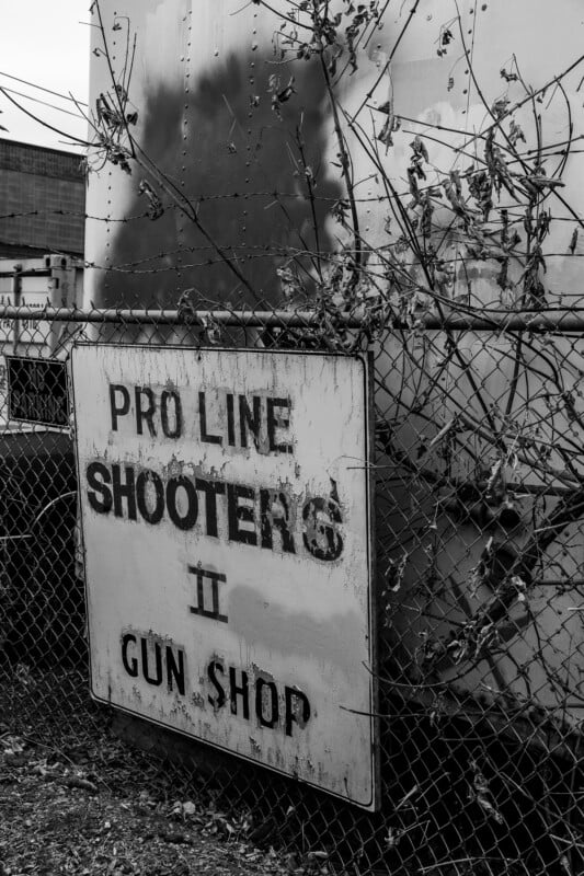 A weathered sign reading "PRO LINE SHOOTERS II GUN SHOP" hangs on a chain-link fence with overgrown vines, in a black-and-white outdoor setting near an industrial building.