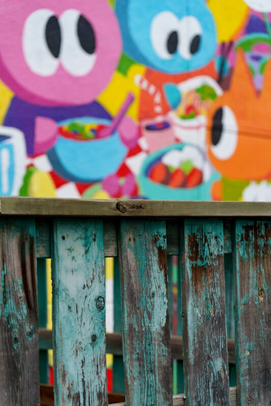 A weathered wooden fence with peeling turquoise paint stands in front of a colorful mural featuring cartoon characters with large eyes, eating food and drinking.