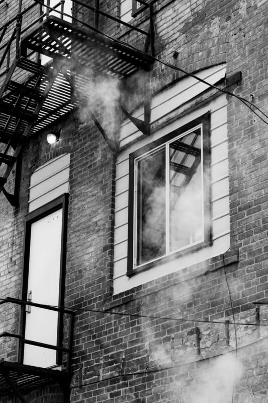Black and white photo of a brick building’s exterior with a fire escape, a door, a window, and steam or smoke rising in front of the structure.