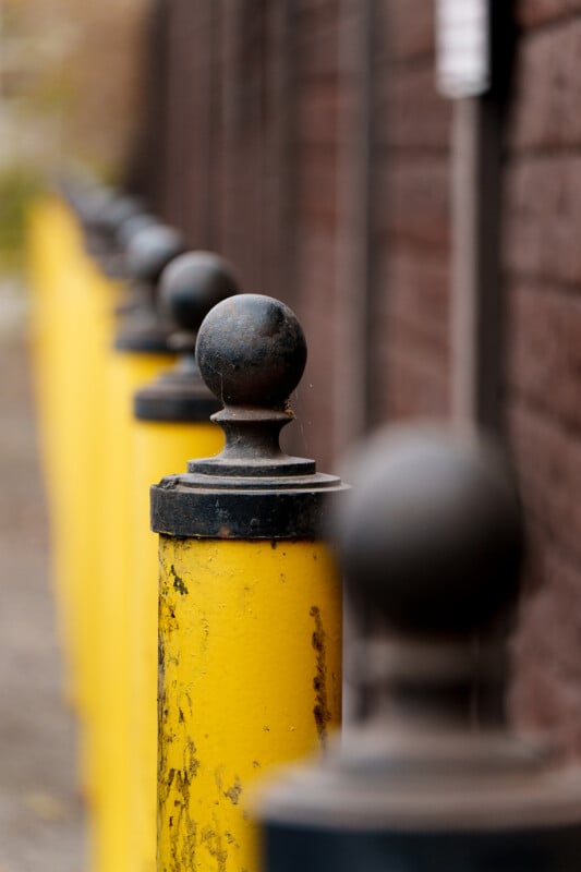A row of yellow metal bollards with round, dark tops lines the edge of a sidewalk next to a brick wall, with the nearest bollard in sharp focus and the others blurred in the background.