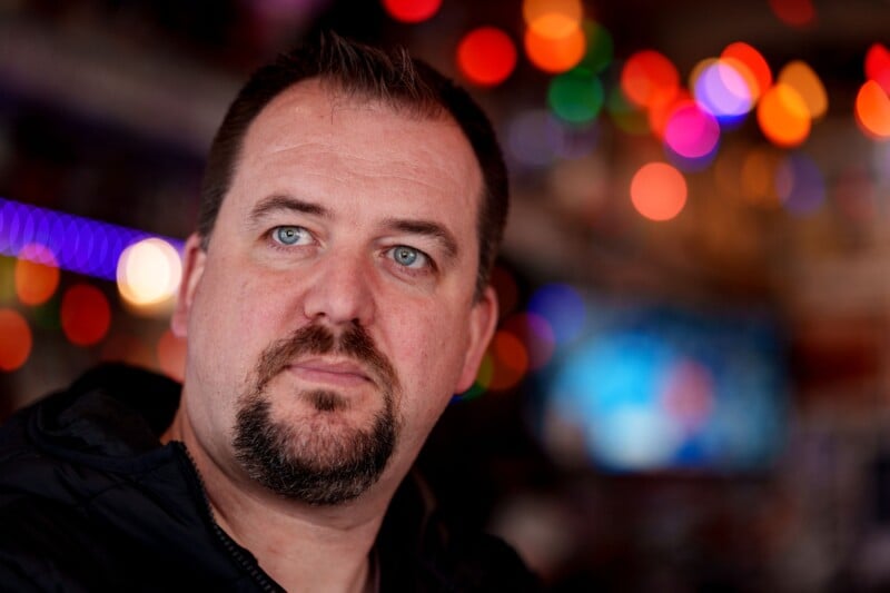 A man with short hair and a goatee looks into the distance. The background is blurred with colorful bokeh lights, creating a warm and vibrant atmosphere. The man is wearing a dark jacket.