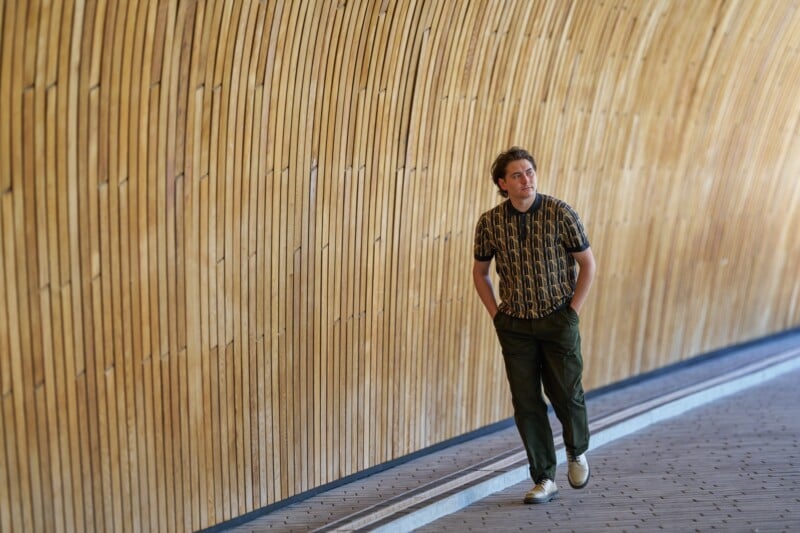 A man with hands in his pockets walks along a curved, wooden wall in a tunnel. He is wearing a patterned shirt, olive green pants, and white sneakers. The scene is brightly lit.