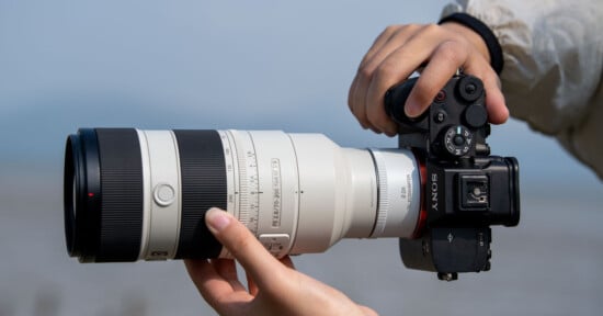 A person holding a Sony camera equipped with a large white telephoto lens, positioned against a blurred outdoor background.