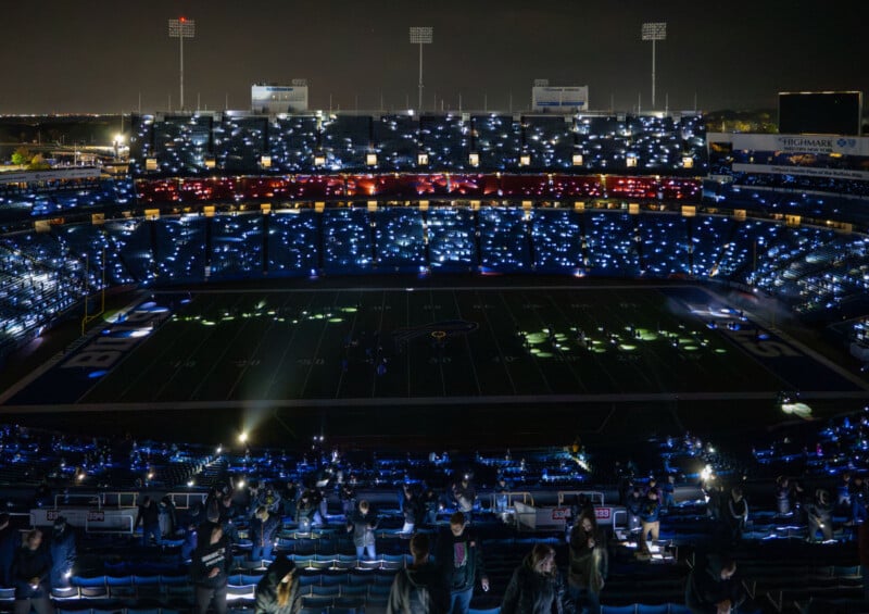 A football stadium at night with the field dark, while thousands of fans in the stands shine lights from their phones, creating a sparkling effect across the arena.