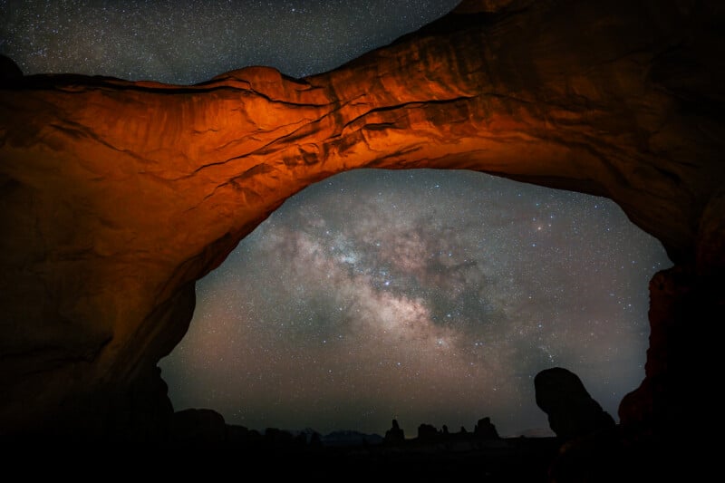 A natural rock arch is silhouetted against a starry night sky with the Milky Way visible. The arch is illuminated with a warm orange light, contrasting with the deep blues and blacks of the sky and distant rock formations.