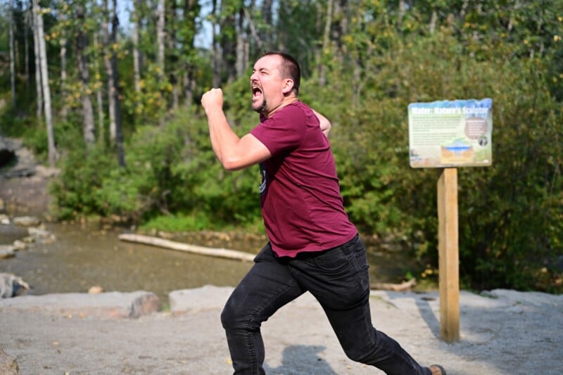 A man with short hair and a beard, wearing a maroon shirt and black pants, runs energetically outdoors near a creek, with trees and a signboard visible in the background.