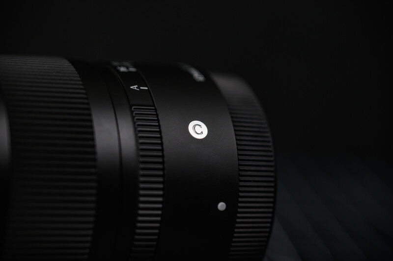 Close-up of a black camera lens with textured focus rings and a white "C" marking, set against a dark, blurred background.