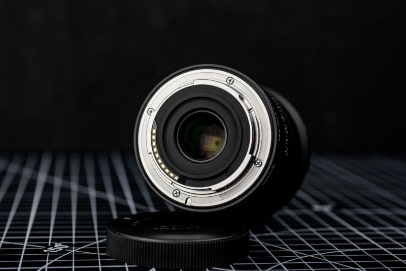 A close-up view of a camera lens lying on its side, showcasing the lens mount and electronic contacts, with a lens cap placed nearby on a black grid-patterned surface.