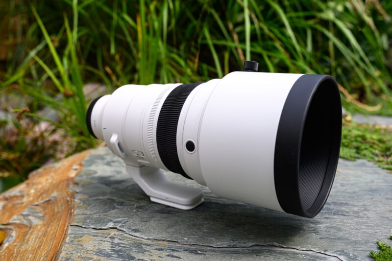 A large white telephoto camera lens rests on a stone surface outdoors, with green grass in the blurred background.