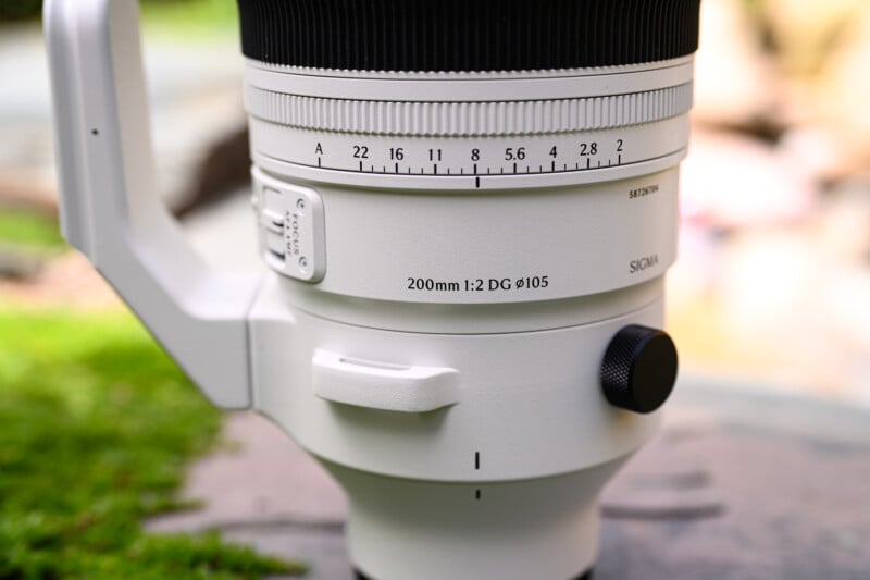 Close-up of a white Sigma camera lens with visible focus and zoom markings, labeled “200mm 1:2 DG ø105.” The lens is resting outdoors on a surface, with blurred grass and natural background.