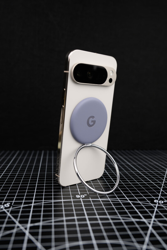 A smartphone with a white case stands upright on a grid-patterned surface. It has a round, lavender Google-branded grip with a metal ring attached to the back. The background is dark and plain.