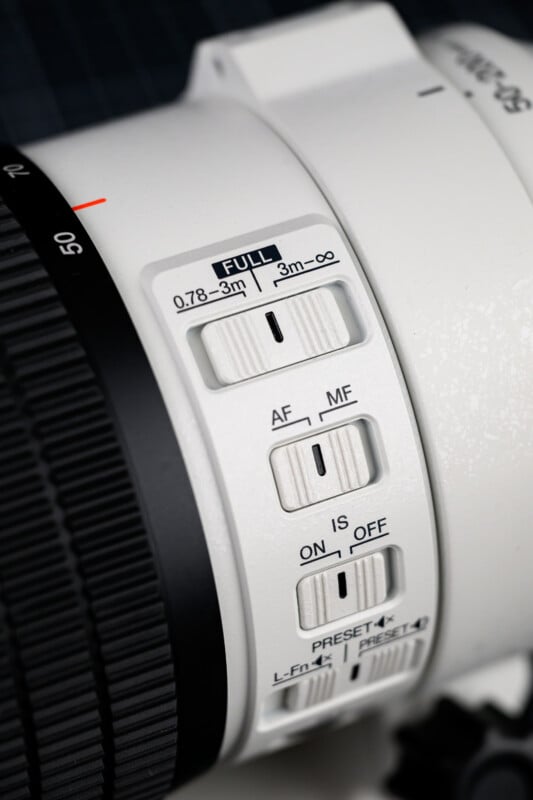 Close-up of switches and dials on a white camera lens, showing focus range, AF/MF, IS on/off, and preset buttons, with black textured focus ring partially visible on the left.