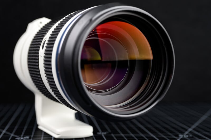 Close-up of a camera telephoto lens with visible reflections on the glass, set on a white tripod mount against a dark background with grid lines.