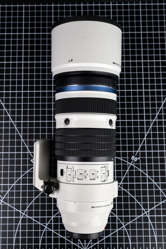A white telephoto camera lens with multiple control switches and a tripod mount, placed vertically on a black gridded surface with white measurement markings.