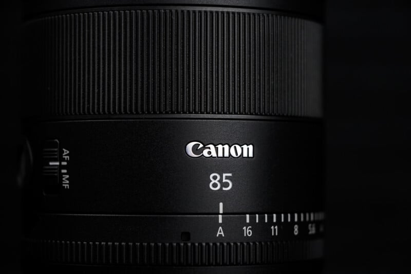 Close-up of a black Canon camera lens with "Canon" and "85" clearly visible, along with focus and aperture markings, on a dark background.