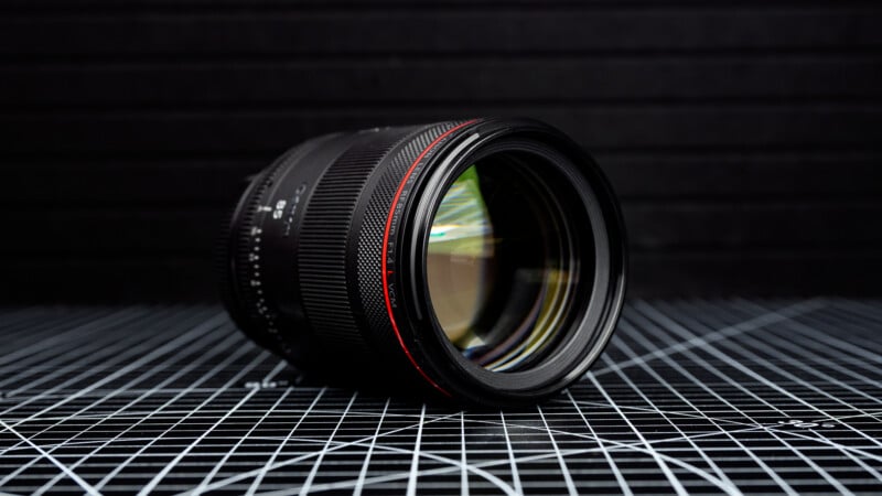 A camera lens with a red ring detail lies on its side on a black and white grid-patterned surface, with a blurred dark background.