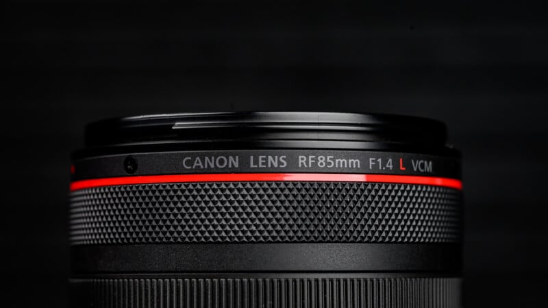 Close-up of a Canon RF 85mm F1.4 L VCM camera lens, showing the textured focus ring and red accent line, with the lens name clearly visible against a dark background.